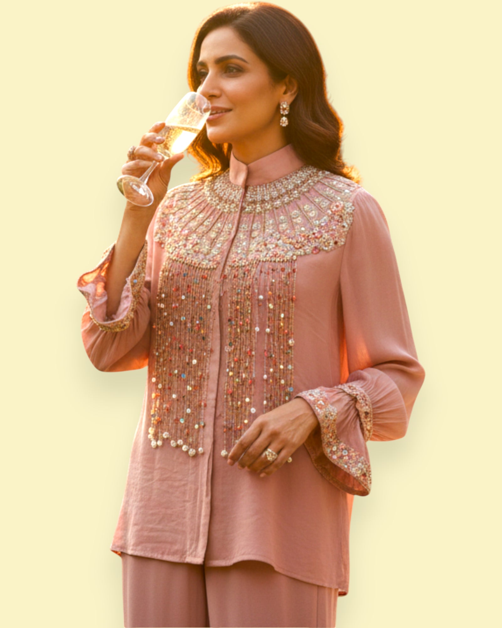 Woman in an embroidered pink traditional outfit holding a glass of champagne against a light yellow background