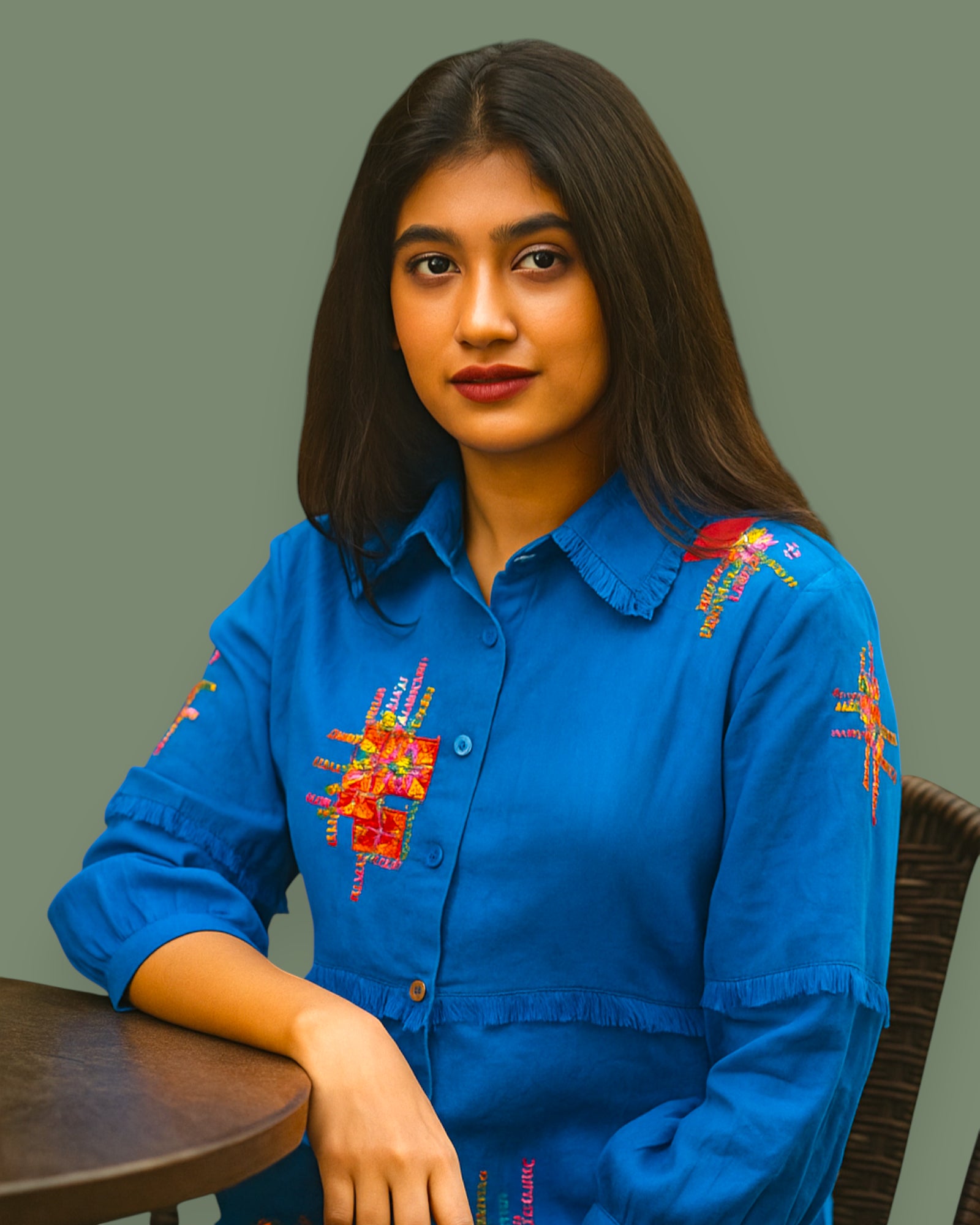 Woman wearing a blue embroidered shirt sitting at a table with a plain background