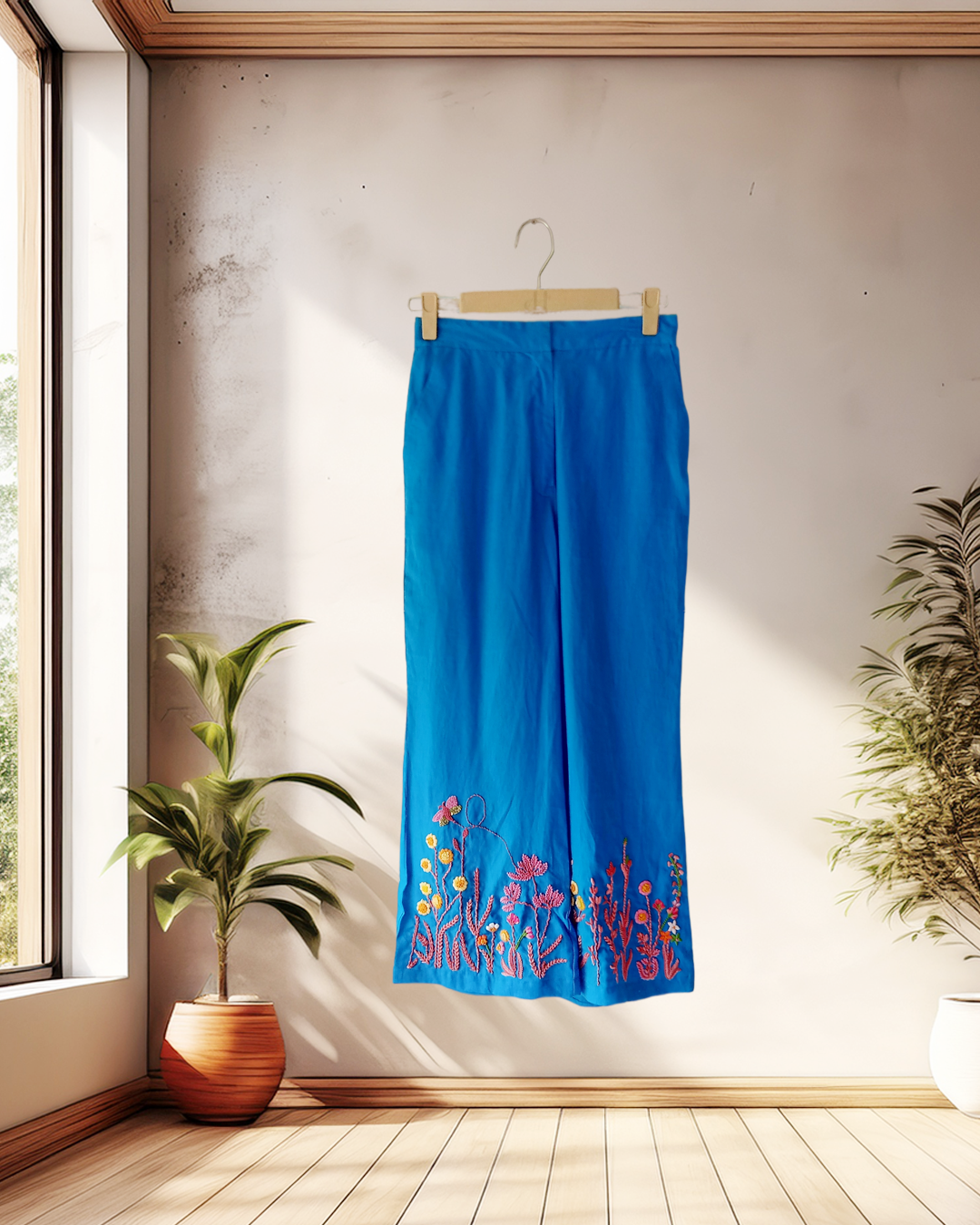 Blue pants with floral embroidery hanging on a wooden hanger against a light wall.