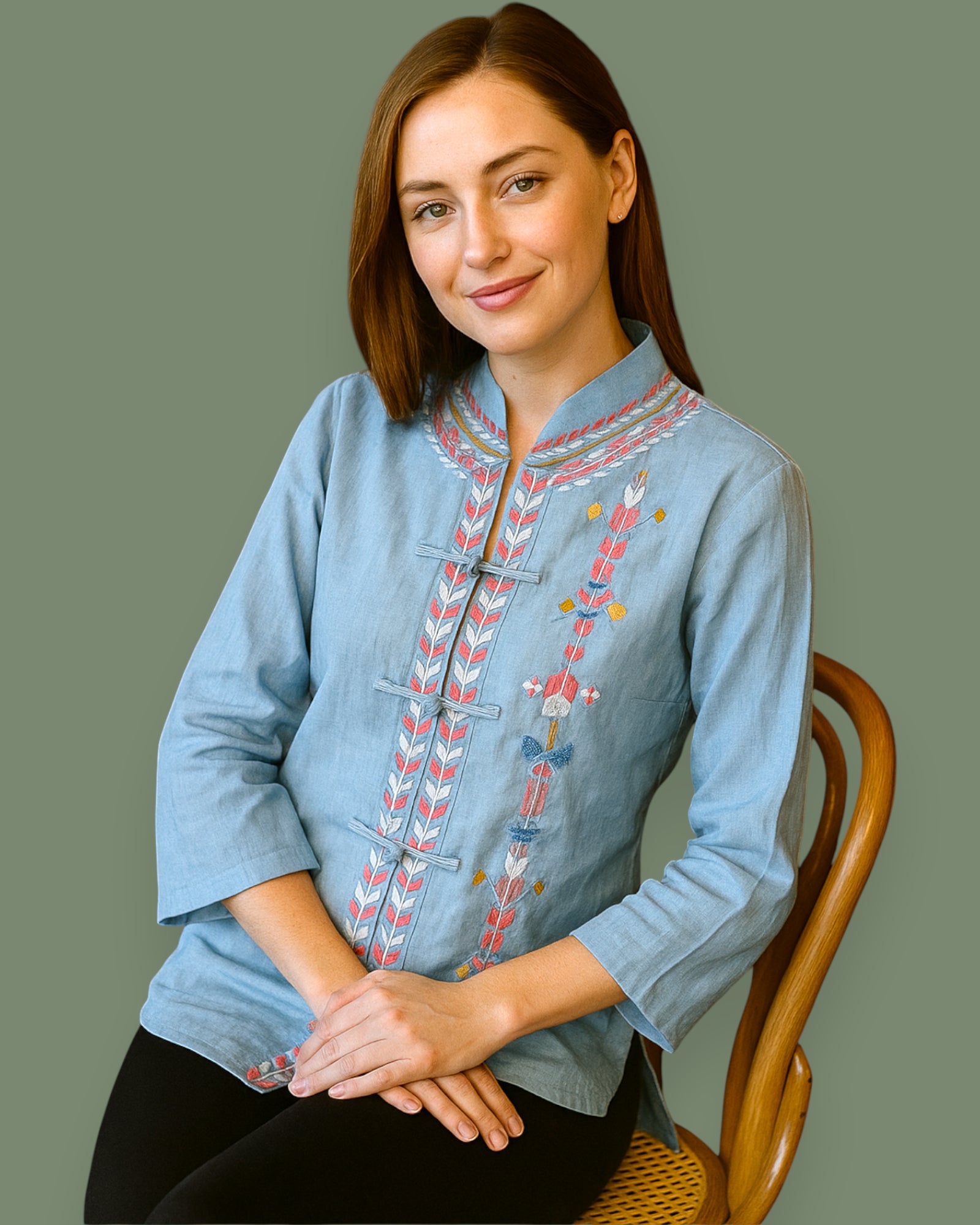 Woman wearing a blue embroidered shirt sitting on a wooden chair against a plain background
