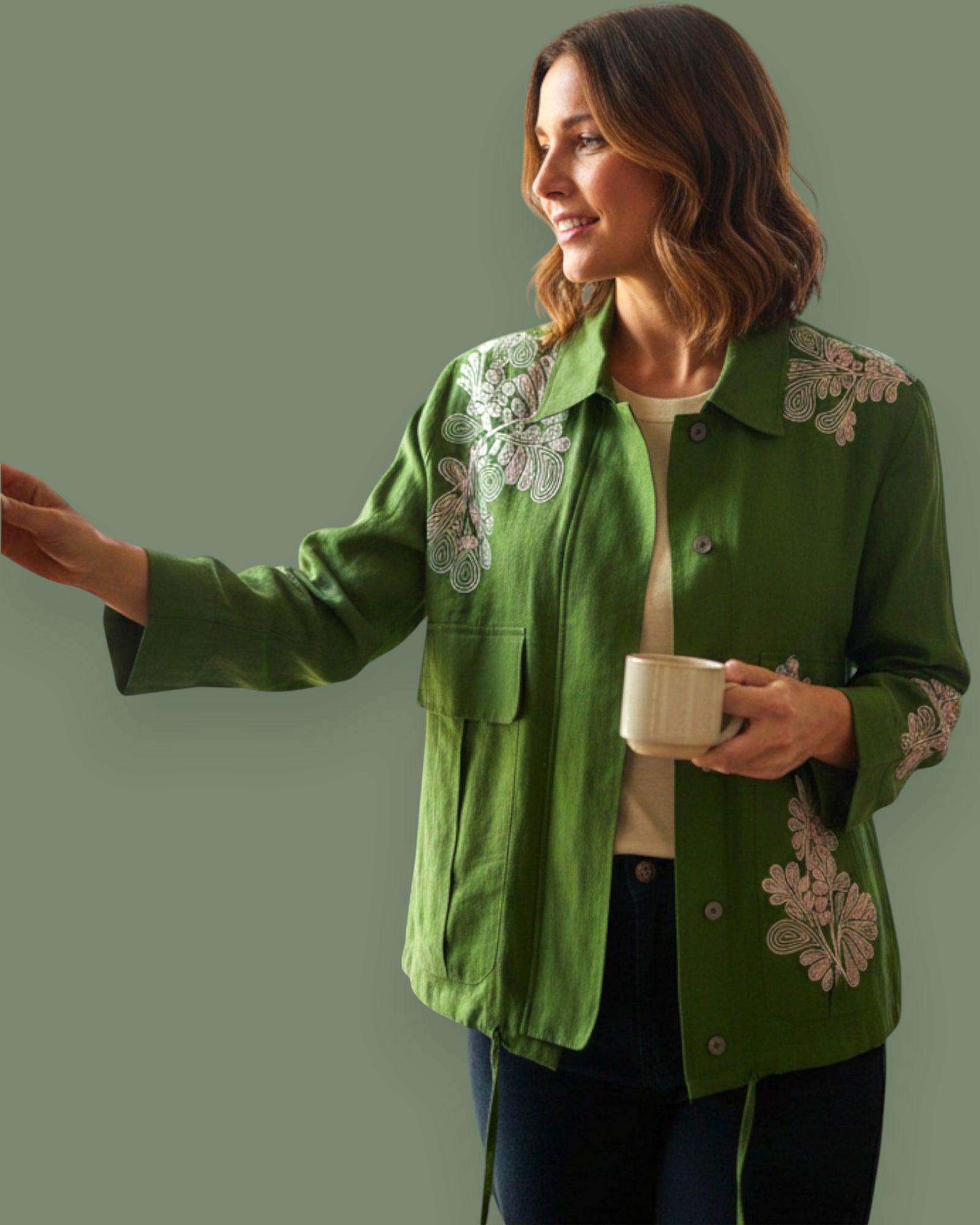 Woman wearing a green jacket with floral embroidery, holding a cup against a plain background