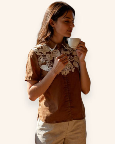 Woman wearing a brown Shirt with lace details, holding a cup against a plain background