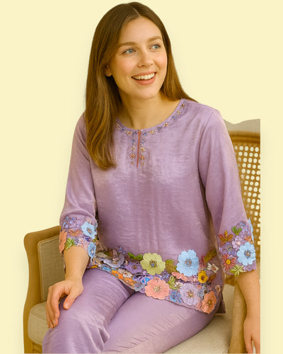 Woman wearing a purple embroidered top sitting on a chair with a plain background