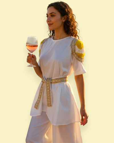 Woman in a white dress with gold accents holding a glass against a yellow background