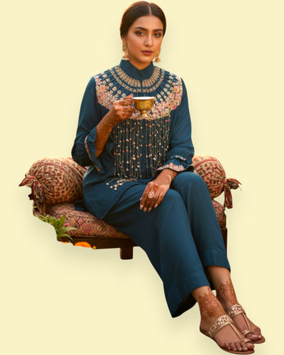 Woman in traditional embroidered outfit sitting on a decorative chair holding a cup.