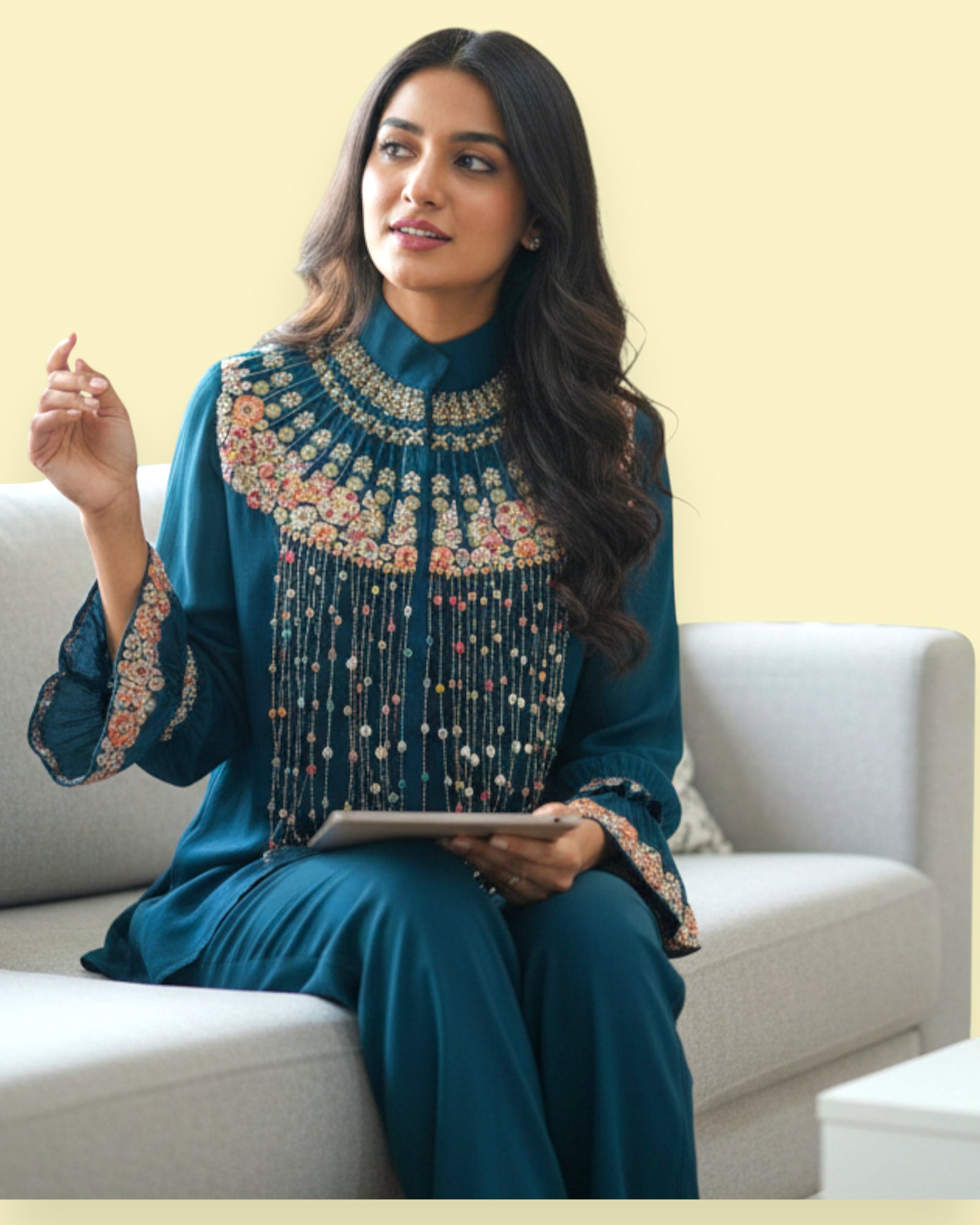 Woman in a teal embroidered outfit sitting on a white couch with a light yellow wall.