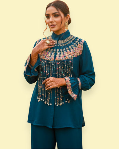 Woman wearing a teal traditional outfit with intricate embroidery on a light yellow background
