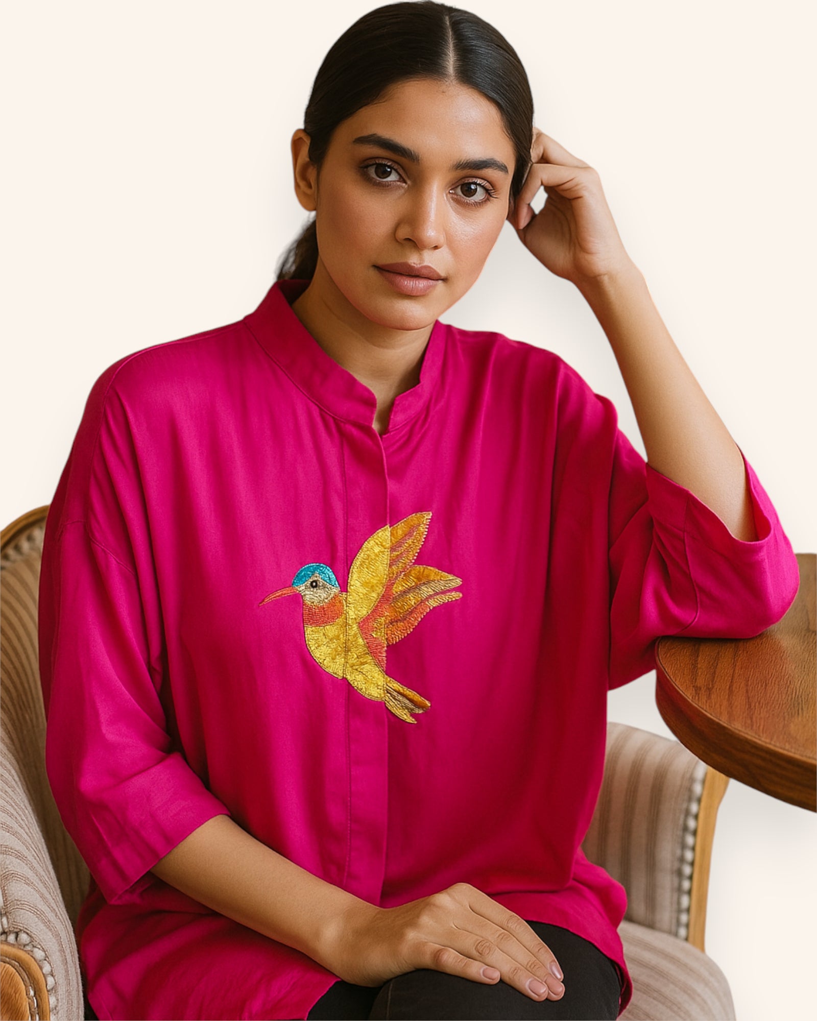 Woman wearing a bright pink kurta with a colorful bird embroidery design, sitting on a chair.