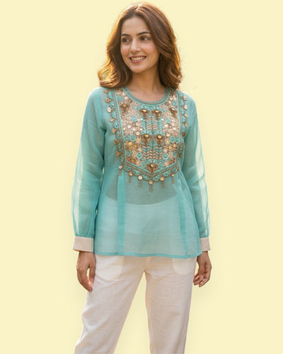 Woman wearing a light blue embroidered top with white pants on a yellow background