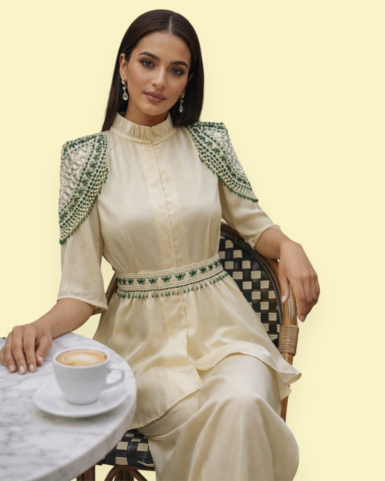Woman in a cream traditional outfit with green patterns sitting next to a coffee cup on a light background