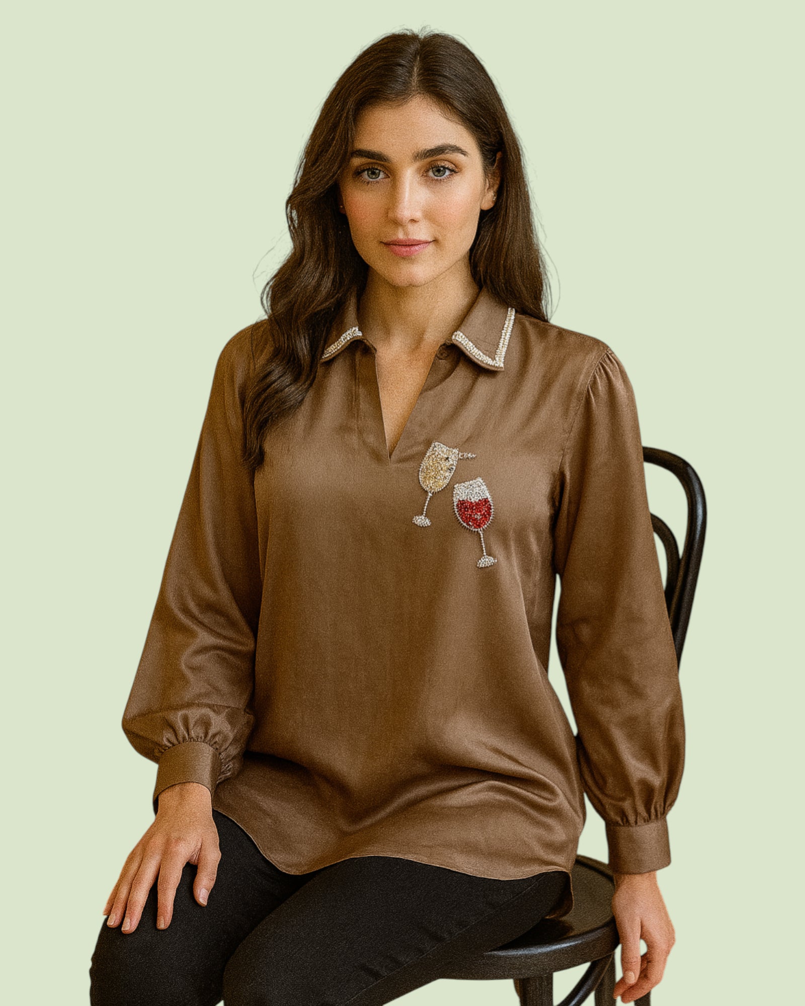 Woman wearing a brown Shirt with wine-themed design sitting on a chair against a light green background
