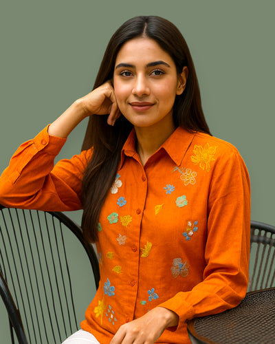 Woman wearing an orange floral embroidery shirt sitting on a chair against a plain background