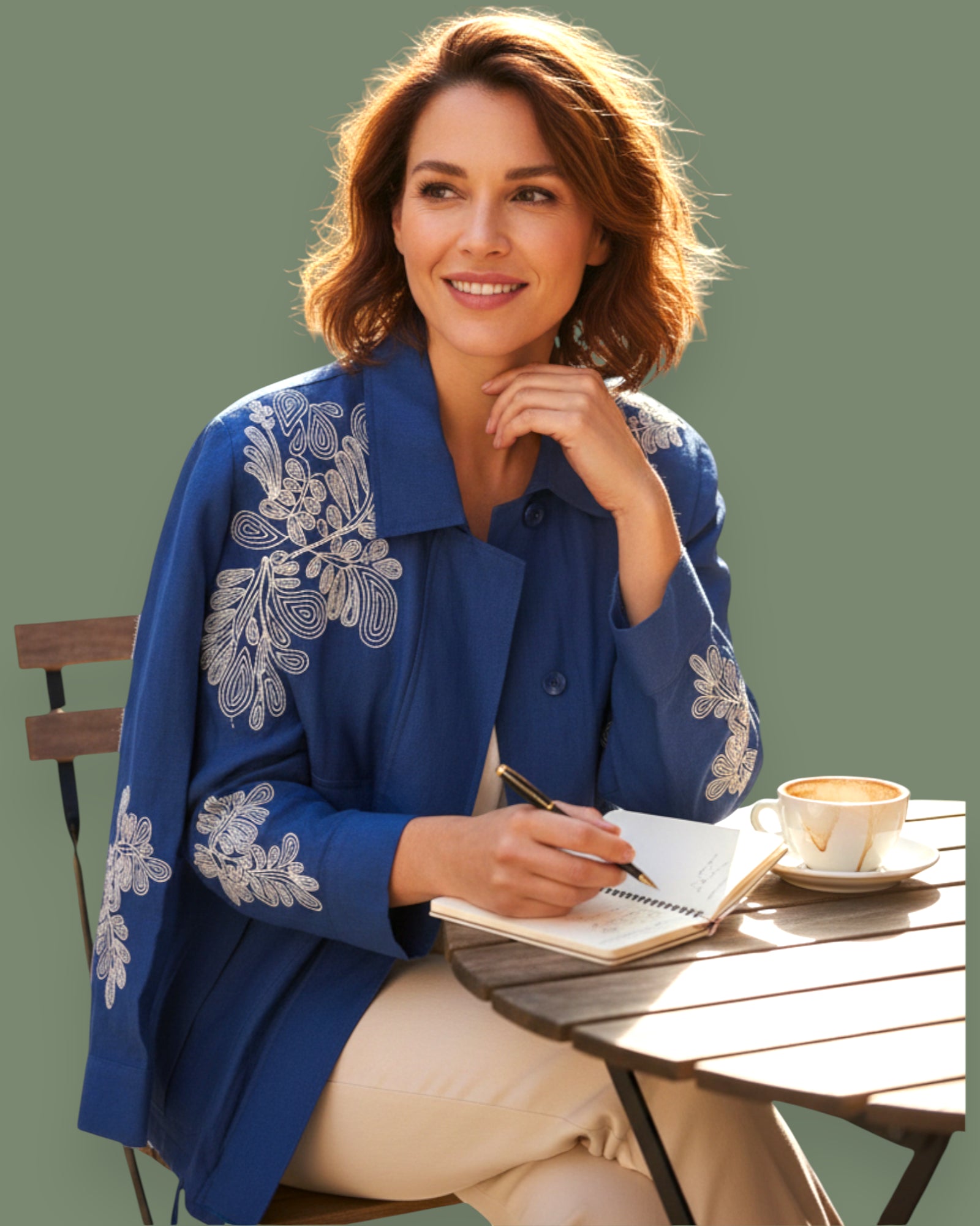 Woman in a blue embroidered jacket sitting at an outdoor table with a notebook and coffee.