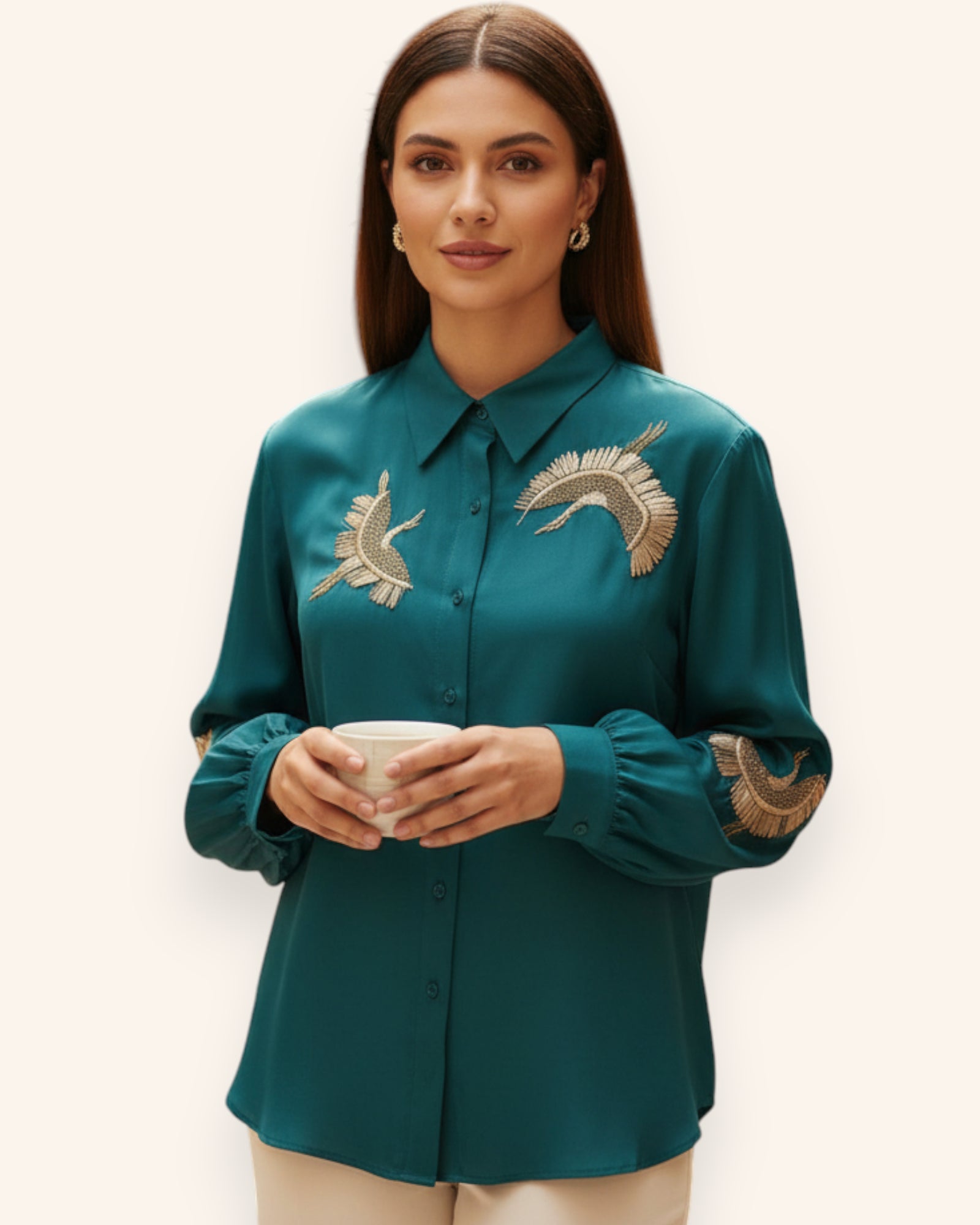 Woman wearing a teal shirt with floral embroidery, holding a cup against a white background